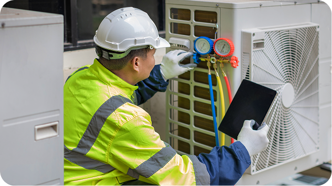An HVAC technician wearing a white hard hat and high-visibility vest uses a tablet while checking pressure gauges on an outdoor air conditioning unit.