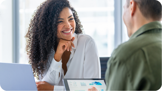 A smiling female tax professional with curly hair sitting at a desk and talking with a male client holding a tablet.