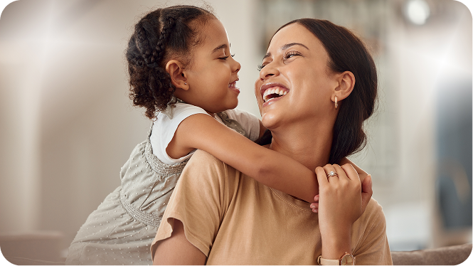 A smiling mother and her young daughter sharing a joyful moment. The daughter, with braided hair, is leaning over her mother's shoulder from behind, hugging her neck, while both laugh together in a warmly lit living room.