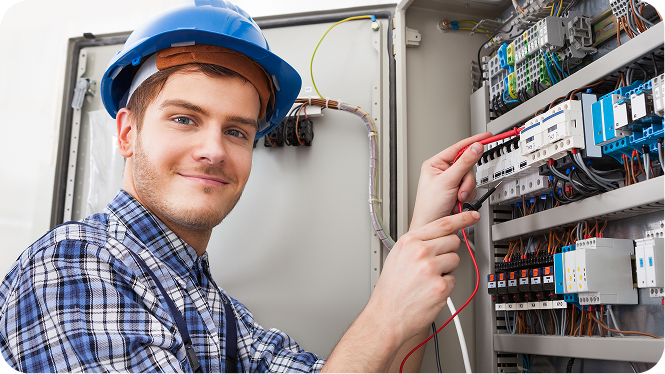 A smiling young male electrician wearing a blue hard hat and plaid shirt uses a digital multimeter to test wiring inside an electrical panel.