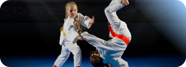 Here is an alt text option tailored for ADA / WCAG compliance for this martial arts image:  Alt Text:  Two young children practicing martial arts on a blue mat, with one child in a defensive stance and the other performing a dynamic mid-air kick.