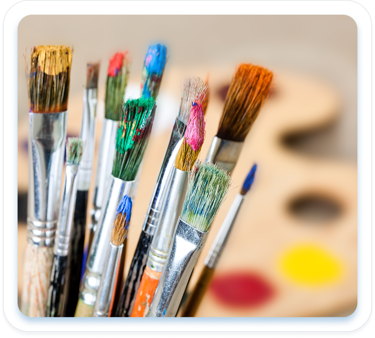 Close-up of a cluster of paintbrushes with bristles covered in blue, green, pink, and orange paint, in front of an artist's wooden paint palette.