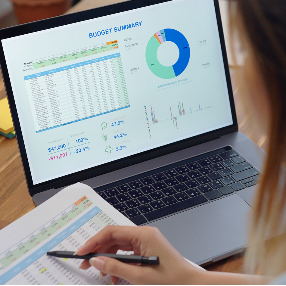 Accountant reviewing budget summary dashboard on laptop displaying financial charts, budget allocations, expense categories, and key performance metrics alongside printed financial statements