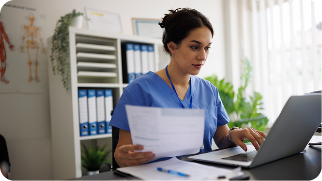 A nursing informatics specialist reviewing paperwork and entering data into a laptop in an office setting.