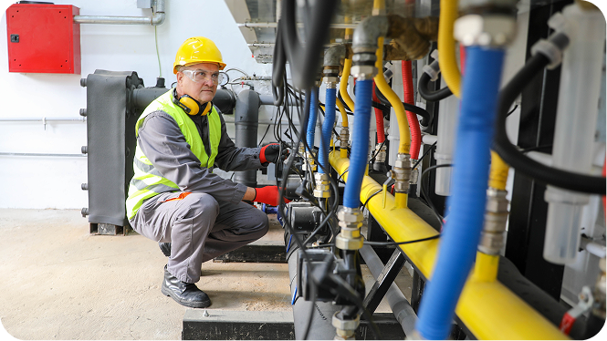 A commercial plumber or technician wearing a yellow hard hat, safety glasses, a high-visibility vest, and protective gloves crouches in an industrial mechanical room, inspecting and working on a complex network of colorful pipes and machinery.