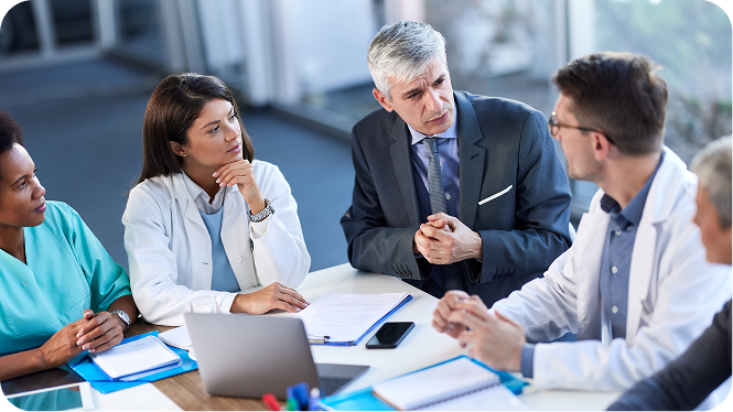 A group of healthcare professionals and an executive in a business suit sitting around a table in a meeting room, reviewing documents and discussing options during a collaborative strategy session.