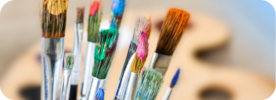 A close-up of several paintbrushes covered in colorful paint with a blurred wooden artist palette in the background
