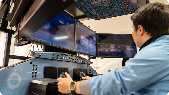 A teenager sitting in a flight simulator cockpit, gripping the controls and looking at multiple overhead and front-facing display screens.