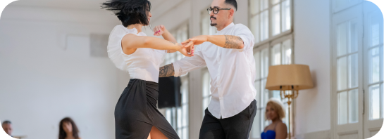 A man and woman holding hands while partner dancing in a bright, sunlit room with large windows.