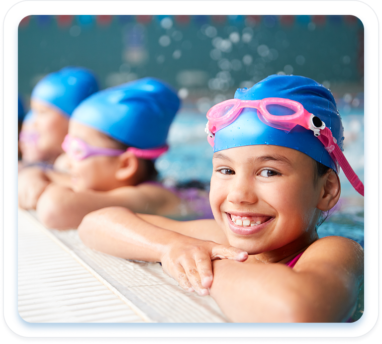 A young girl smiling at the side of a swimming pool, wearing a blue swim cap and pink goggles resting on her forehead, with other children lined up behind her.