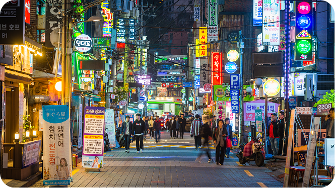 A vibrant, brightly lit street in South Korea at night, lined with dense, glowing neon and LED signs in Korean characters. Pedestrians walk down the center of the paved road, which features a painted white directional arrow pointing forward. Upright banner stands and storefronts line both sides of the street under a complex canopy of overhead power lines.