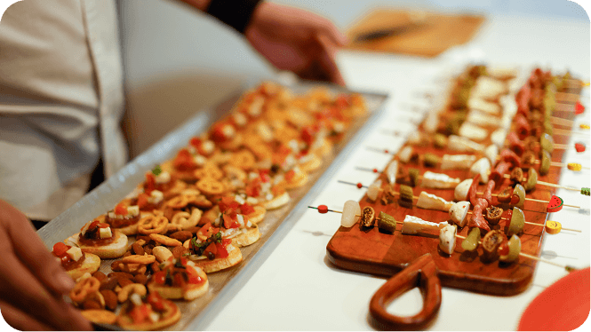 Server presenting a tray of bite-sized appetizers, including bruschetta and skewered hors d'oeuvres, on a catering table.