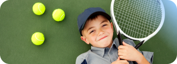 A young boy wearing a cap lies on a green tennis court next to three tennis balls, holding a tennis racket across his chest and smiling at the camera.