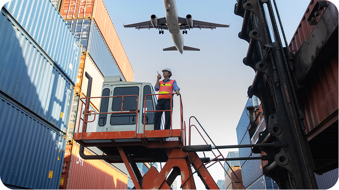 A logistics worker wearing a safety vest and hard hat stands on an elevated platform at a shipping port, holding a walkie-talkie as a cargo plane flies overhead between stacked shipping containers.