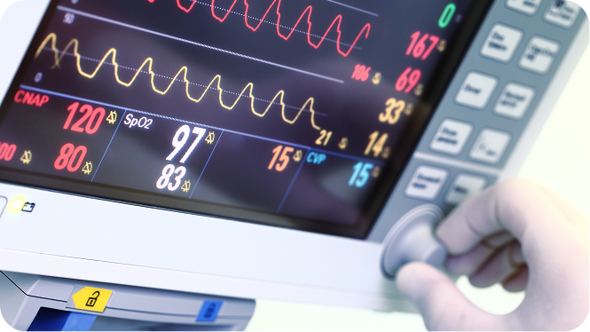 Close-up of a medical monitor displaying real-time patient vitals—including a continuous heart rhythm wave, an SpO2 reading of 97%, and a blood pressure reading of 120 over 80—as a gloved hand adjusts a control dial on the right side.