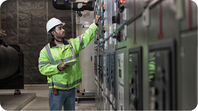 A commercial electrician wearing a white hard hat and high-visibility jacket inspecting a large industrial electrical control panel while holding a digital tablet.