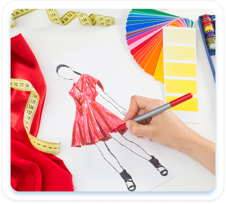 A hand sketches and colors a red dress design on paper, surrounded by red fabric, a measuring tape, and a fan of colorful marker swatches.