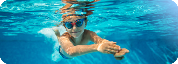 A young boy wearing blue goggles swimming underwater in a bright blue pool, extending his arms forward toward the camera.