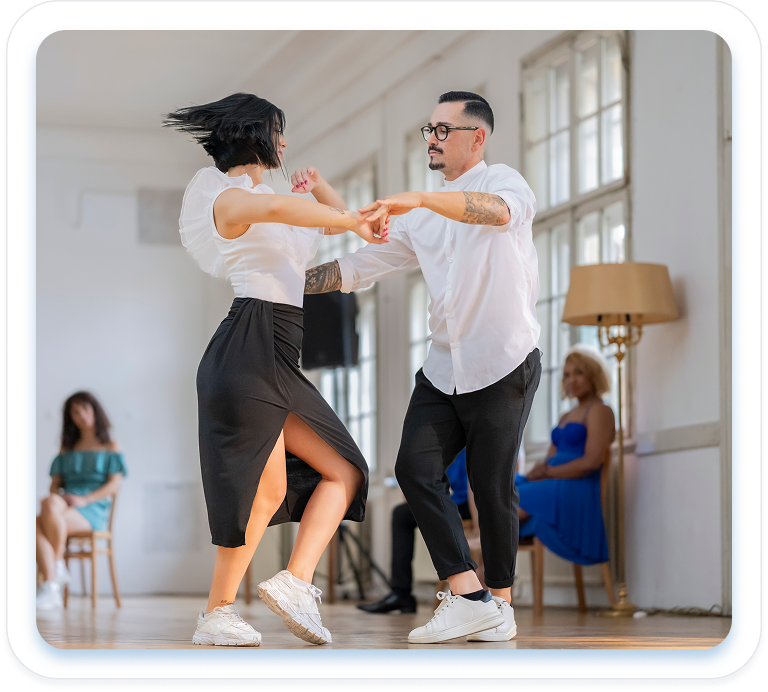 A man and woman in white tops and black outfits dance together in a bright, sunlit studio while onlookers watch from chairs in the background.
