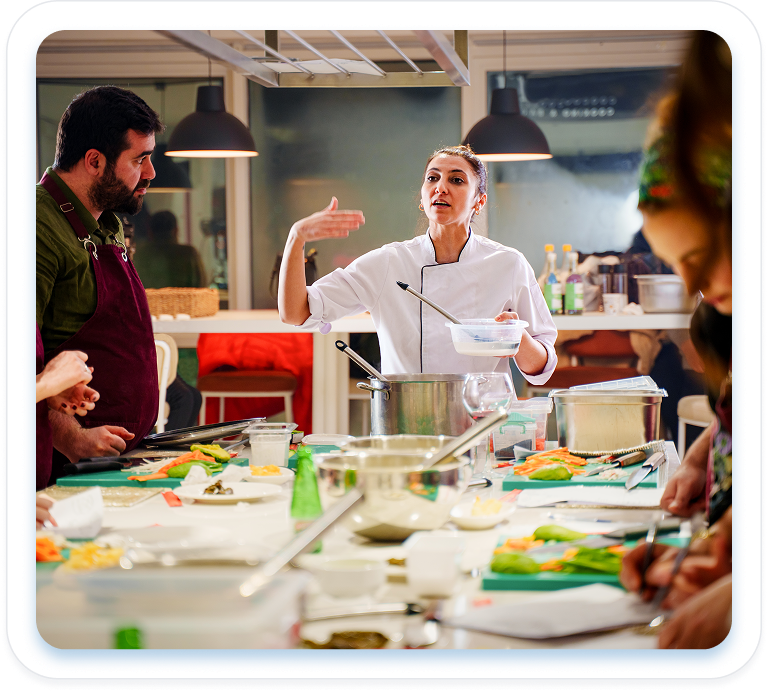 Female chef instructor in a white uniform gesturing while teaching a cooking class to adults gathered around a kitchen prep table filled with ingredients."