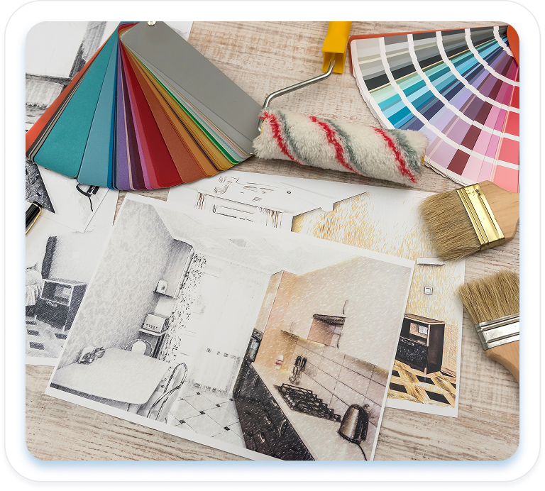 A tabletop covered with interior design and craft materials: fanned-out color swatch samples, a paint roller, hand-drawn room sketches, and two paintbrushes.