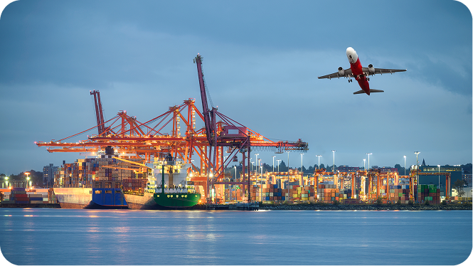 A bustling commercial shipping port at dusk or dawn, featuring large orange container cranes loading cargo ships docked along the water. In the upper right sky, a commercial airplane with a red and white belly is taking off or flying low over the harbor.