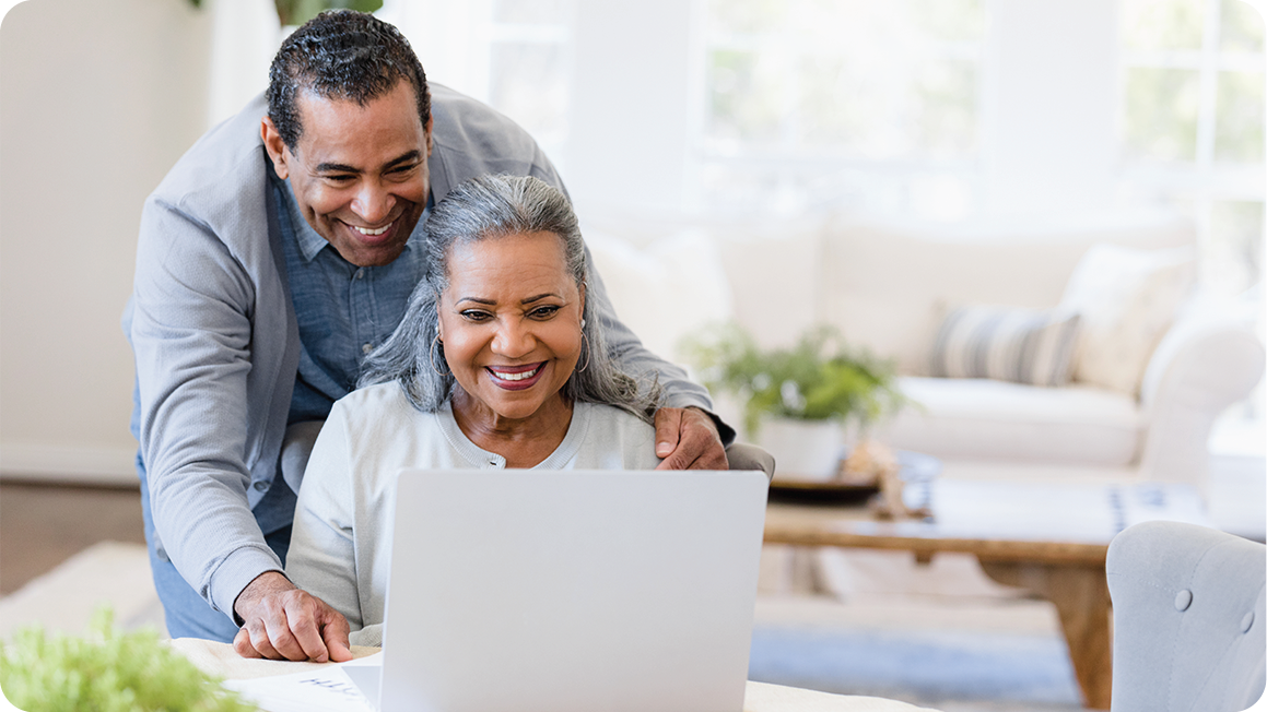 An older woman with silver-gray hair and a white top sits at a laptop, smiling warmly, while a younger man in a light gray sweater and blue shirt stands beside her, leaning in and laughing. They are in a bright, modern living room with a white sofa, wooden coffee table, and green plants, creating a warm and supportive atmosphere. The pair appear engaged and happy while working together on the laptop, suggesting a close familial or mentoring relationship. Soft natural light filters through windows, highlighting their expressions of joy and connection.