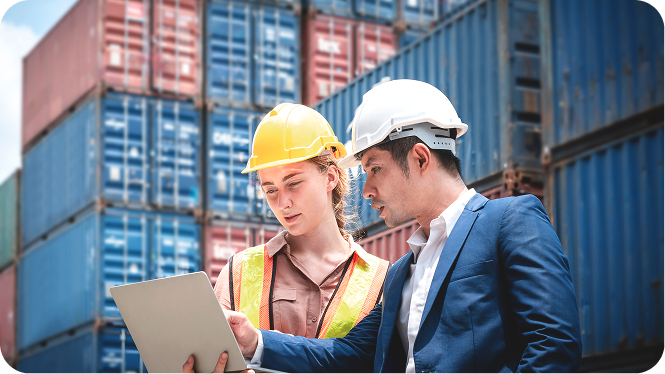 A female site inspector in a yellow hard hat and safety vest and a male logistics manager in a white hard hat and suit looking at a laptop computer in a shipping container yard.