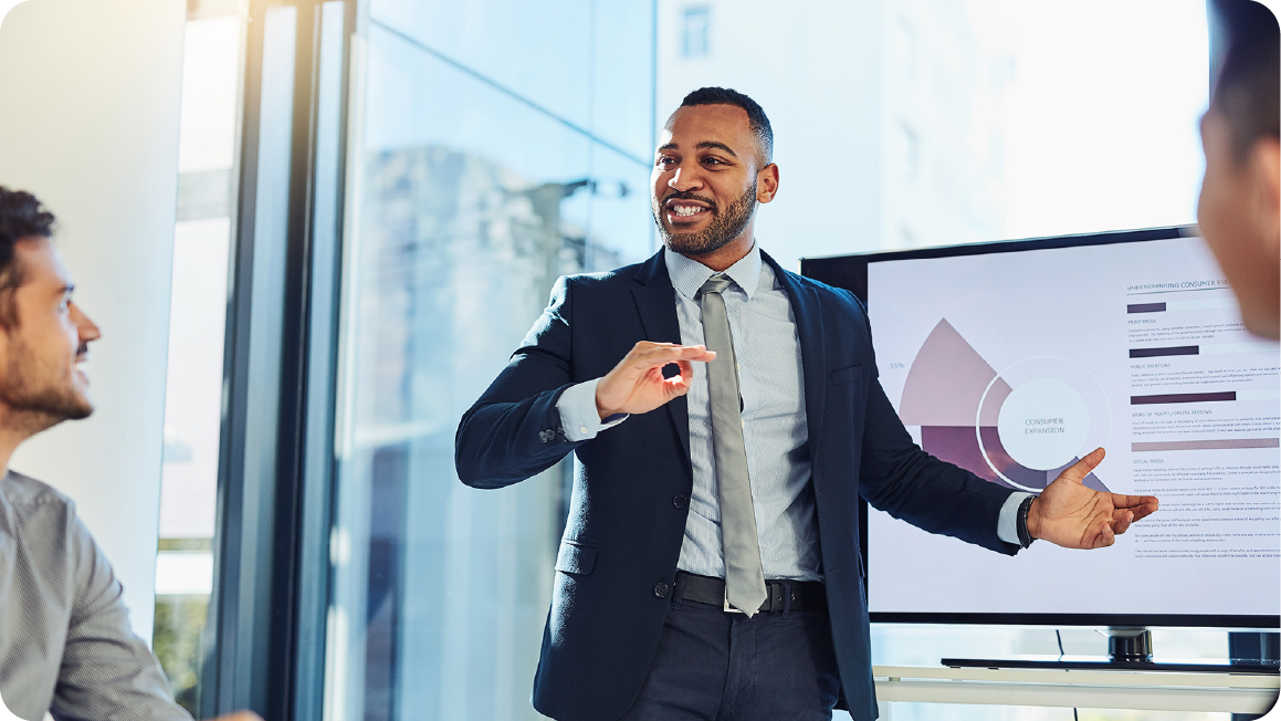 Business professional in a navy suit and gray tie presenting a data visualization on a screen to colleagues in a modern, bright office setting. He stands confidently, smiling and gesturing with an open hand towards a purple and white pie chart or segmented circular graphic. The presenter appears engaged and enthusiastic, with soft natural light filtering through large windows behind him. Another professional is partially visible in the foreground, listening attentively. The sleek environment suggests a corporate meeting or strategy discussion focused on analytics and business performance.