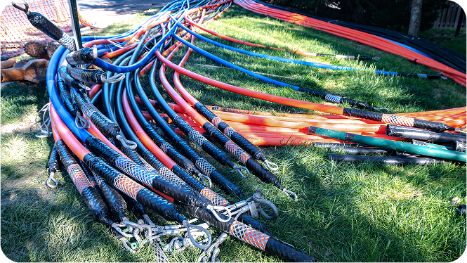 A bundle of orange, blue, and green fiber optic conduits fitted with mesh pulling grips and shackles, laid out on green grass for underground installation or cable blowing.