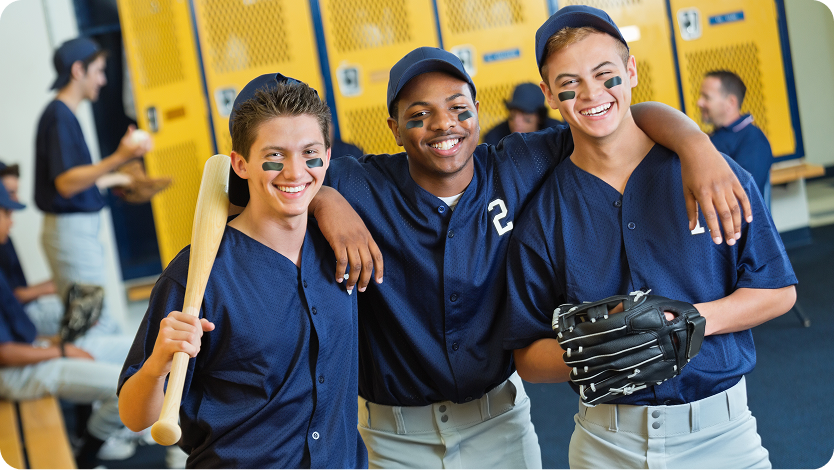 Three smiling teenage baseball players in blue uniforms and caps pose together with their arms around each other in a locker room. One holds a wooden bat, and another holds a catcher's mitt.