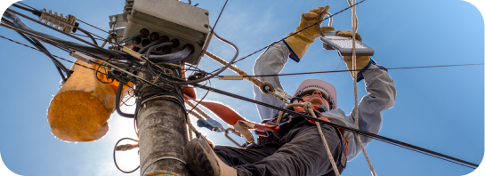 An electrician in safety gear, including a hard hat and gloves, working on a utility pole with many wires against a bright blue sky, representing.