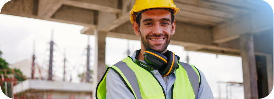Smiling construction worker in a yellow hard hat and safety vest, with ear protection, representing opportunities in Construction and Trades.
