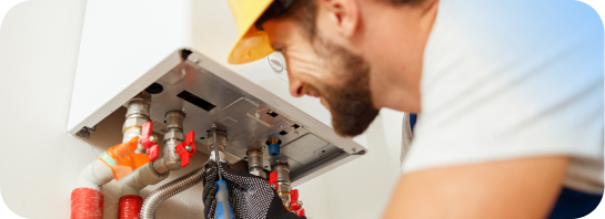 A smiling male technician in a hard hat and gloves uses a screwdriver to work on the pipes of a water heater, representing hands-on training in.