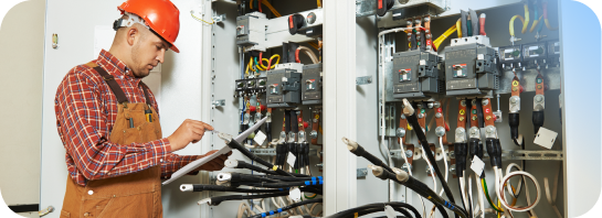 An electrician in a hard hat and overalls inspects wiring inside an open electrical panel, representing hands-on training for Construction and.