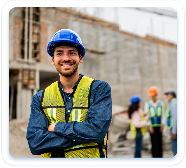 Smiling construction worker in a blue hard hat and safety vest, arms crossed, with colleagues and a building site in the background.