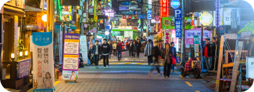 A vibrant, brightly lit street in South Korea at night, lined with dense, glowing neon and LED signs in Korean characters. Pedestrians walk down the center of the paved road, which features a painted white directional arrow pointing forward. Upright banner stands and storefronts line both sides of the street under a complex canopy of overhead power lines.