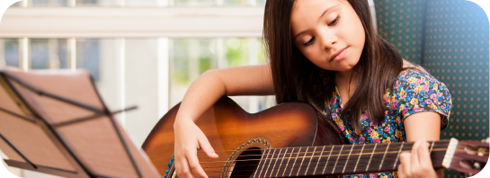 Young girl with long dark hair, wearing a floral shirt, intently playing an acoustic guitar with sheet music nearby.