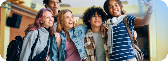 Diverse group of five smiling young adults, some with backpacks, taking a selfie together in a bright hallway.