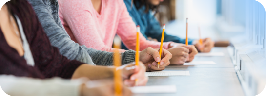 Students in a classroom, seen from the side, writing on papers with pencils, focused on their work.