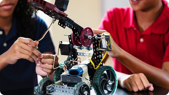 Close-up of hands assembling a technical robotic device, featuring metallic gears, mechanical components, and wheels. Two people partially visible, one wearing a red polo shirt and another in a dark top, collaboratively working on a complex robotics project. The robot appears to be constructed from modular metal and plastic pieces, with blue and yellow structural elements. Hands are carefully manipulating and connecting mechanical parts, suggesting a precise and focused engineering or educational activity, likely part of a robotics workshop or summer camp engineering program.