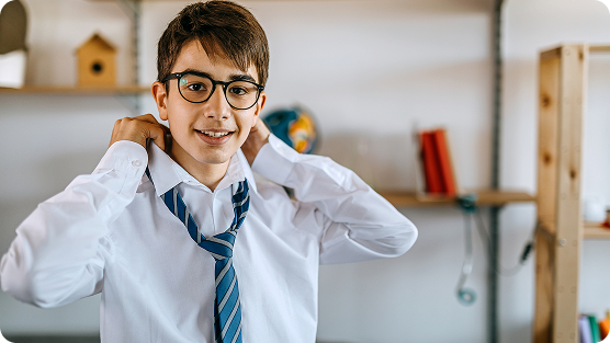 A young student wearing glasses, a white button-up shirt, and a blue-striped tie, stands in a bright classroom or study space with shelves, a birdhouse, and books in the background. He adjusts his tie with a cheerful, confident smile, appearing comfortable and eager. Soft natural lighting illuminates the scene, creating a warm and positive educational atmosphere. The student's posture and expression suggest enthusiasm and readiness for learning.