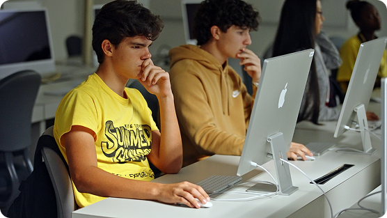 Two students sit intently at white Apple desktop computers in a bright, minimalist computer lab. On the left, a young person wears a bright yellow