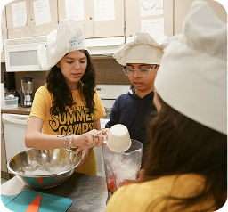 A culinary classroom scene with two students wearing white chef hats, working together at a kitchen counter. A person in a yellow T-shirt with 
