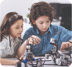 Two young students collaborate intently on a robotic project, seated at a table with mechanical components and electronic devices. They wear casual clothing - one in a blue checkered button-up shirt, the other in a light gray top. Both have focused expressions as they work closely together, leaning over the technical setup. Soft lighting and a blurred background suggest a classroom or workshop setting, highlighting their concentrated teamwork and hands-on learning in what appears to be a science or engineering activity.