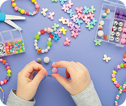 Overhead view of a jewelry-making craft activity on a blue background. Two hands in gray sleeves are threading colorful beads onto a bracelet. Scattered around are bright plastic beads in pastel and primary colors - including flower-shaped and round beads. Transparent storage containers with compartmentalized sections hold additional beads in various sizes and colors. Scissors, partially completed beaded bracelets, and loose beads are spread across the workspace, suggesting a creative and engaging crafting session with a playful, colorful aesthetic.