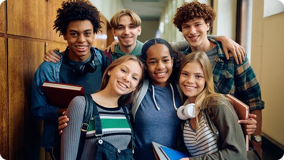 Six diverse high school students grouped together in a school hallway, smiling and looking friendly. A mix of ethnicities and genders, they wear casual clothing like striped and plaid shirts, sweaters, and a blue hoodie. Some carry books and have headphones around their necks. The group stands close, with arms around each other's shoulders, projecting a sense of camaraderie and friendship. The background shows wooden lockers and neutral-toned walls, typical of an educational setting. Their expressions are warm, relaxed, and positive, suggesting a comfortable and supportive peer environment.