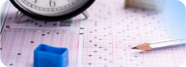 Standardized test answer sheet with pink grid background, partially obscured by a classic analog clock showing time near the top left. A sharp yellow No. 2 pencil rests diagonally on the right side of the sheet, its point positioned ready to mark answers. A solid blue rectangular eraser sits on the left side, creating visual contrast against the pastel pink document. The scene suggests an academic testing environment, with precise instruments and clean, organized layout indicating a formal assessment setting.