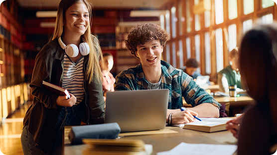 Two students studying together in a bright, sunlit library. A young woman with long hair and white headphones stands holding a book and tablet, wearing a striped top and dark jacket. Beside her, a curly-haired young man sits at a wooden table with an open laptop, writing in a notebook. Wooden bookshelves and large windows create a warm, scholarly atmosphere with soft golden light streaming across the space. Other students can be seen blurred in the background, suggesting a collaborative academic environment.