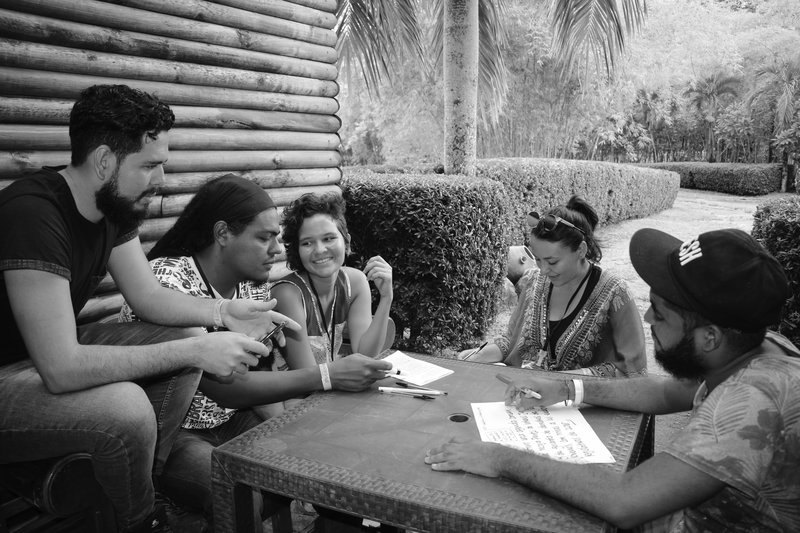 Um grupo de pessoas está ao redor de uma mesa ao ar livre. Elas estão segurando papéis e conversando.